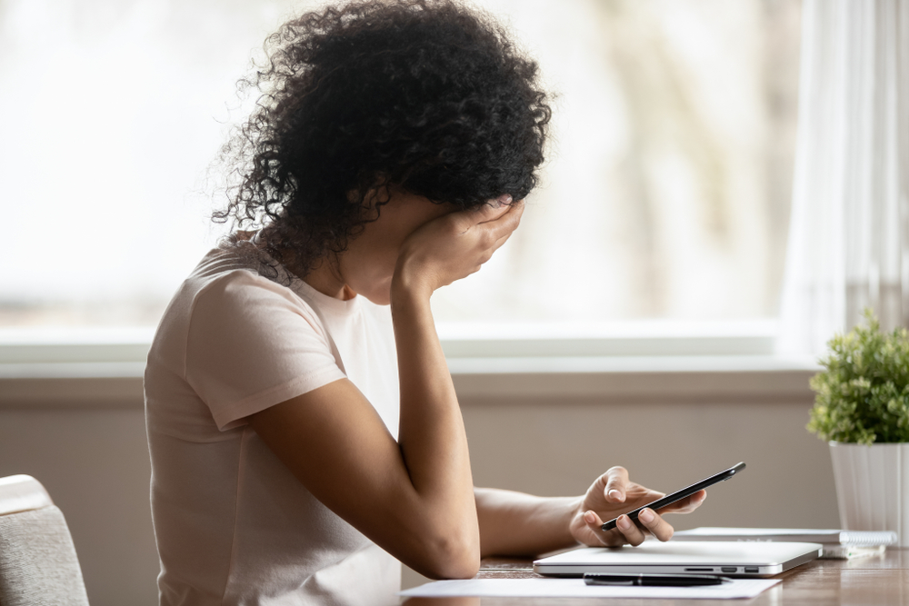 Women sitting at desk with one hand covering her face and the other holding her phone.