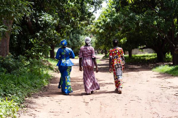 Three women in traditional African dress walk away from camera, down long road, leafy green trees around them