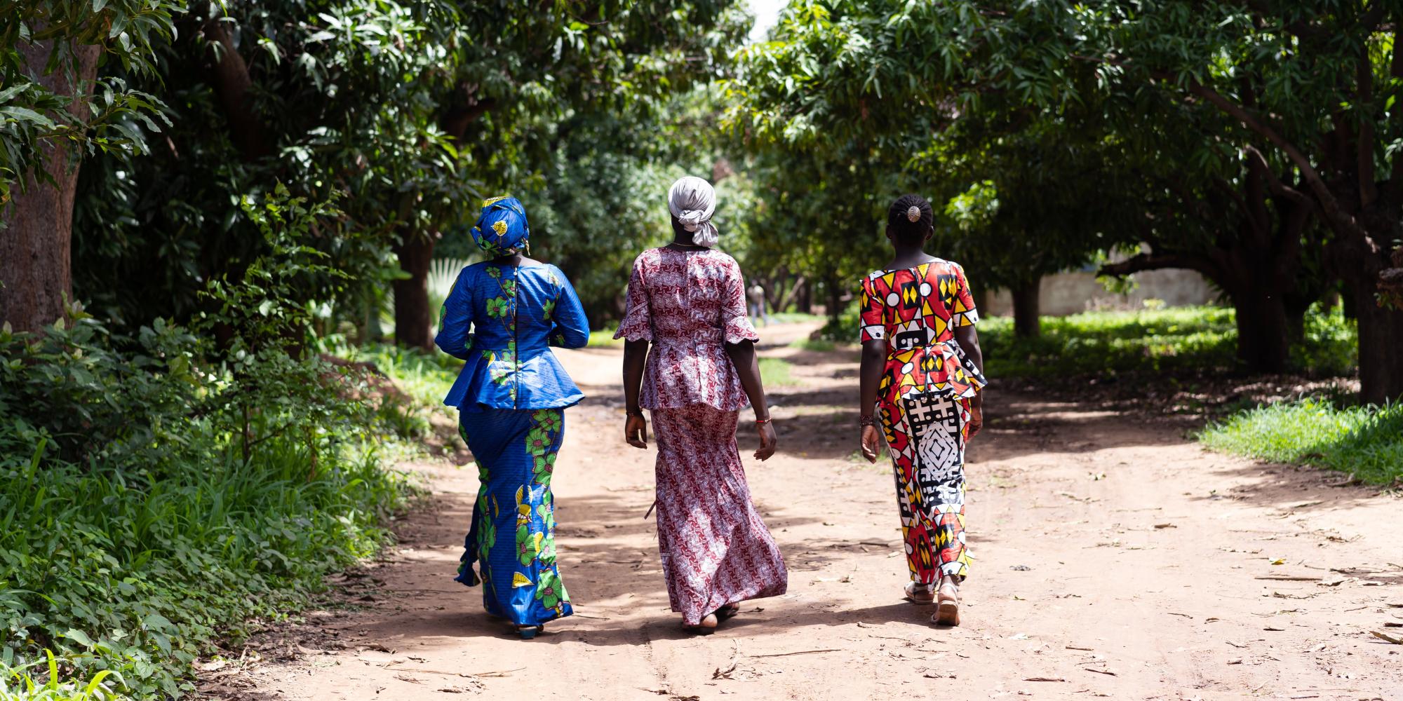 Three women in traditional African dress walk away from camera, down long road, leafy green trees around them